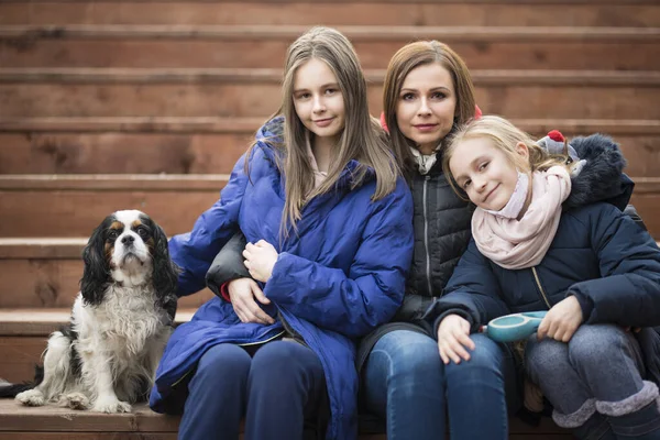 Mom with daughters and a dog are sitting on a wooden staircase