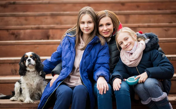 Mom with daughters and a dog are sitting on a wooden staircase