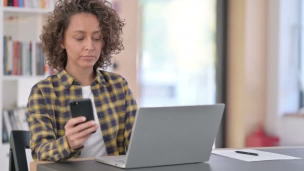 Jeune femme de race mixte utilisant Smartphone et ordinateur portable au travail — Video