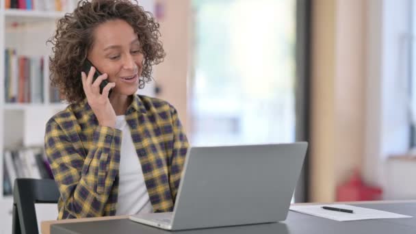 Mujer de raza mixta con ordenador portátil hablando en el teléfono inteligente en el trabajo — Vídeos de Stock