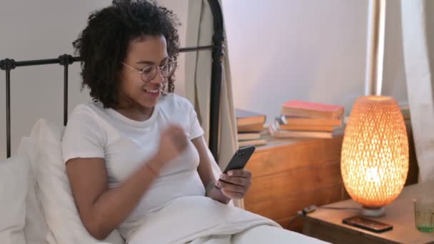 Éxito, Joven mujer africana celebrando en el Smartphone en la cama — Vídeo de stock