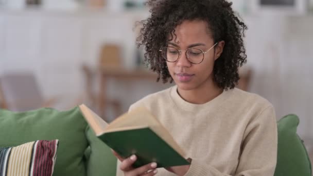 Портрет молодой африканской женщины, сидящей за чтением книги на диване — стоковое видео