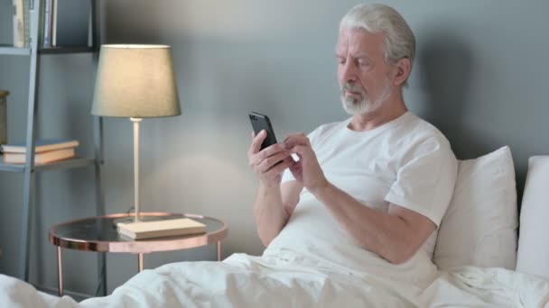 Erfolg: Älterer Mann feiert mit Smartphone im Bett — Stockvideo