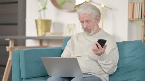 Старший старик с ноутбуком разговаривает на смартфоне на диване — стоковое видео