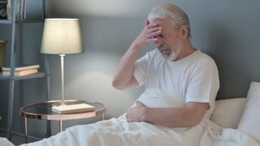 Başağrısı olan yaşlı adam yatakta oturuyor. 