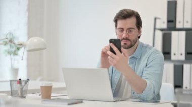 Dizüstü bilgisayarı olan genç adam iş yerinde akıllı telefon kullanıyor.