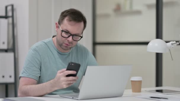 Jonge man met laptop met behulp van smartphone op het werk — Stockvideo