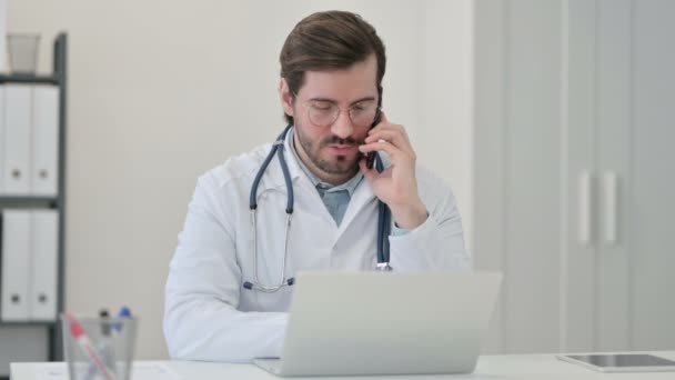 Молодой врач с ноутбуком разговаривает на смартфоне — стоковое видео