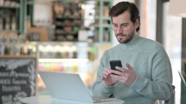 Laptop 'u olan genç adam Cafe' de akıllı telefonu kontrol ediyor.
