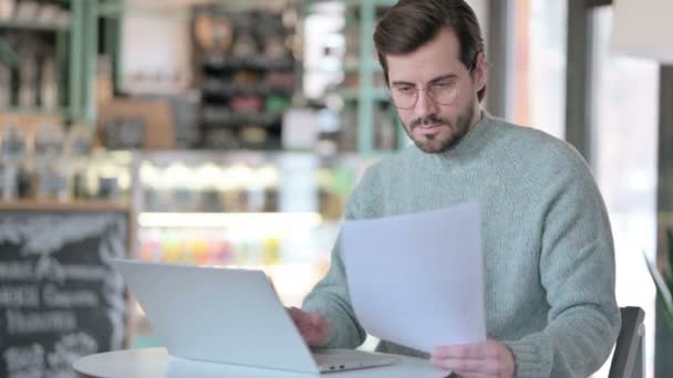 Młody człowiek czytanie papieru podczas korzystania z laptopa w Cafe — Wideo stockowe