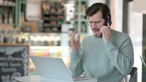 Dühös fiatalember laptoppal beszél okostelefonon — Stock videók