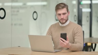 Dizüstü bilgisayar üzerinde çalışırken akıllı telefon kullanan yaratıcı genç adam