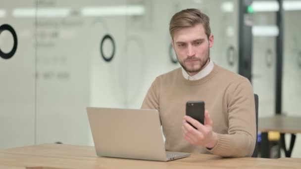 Kreativer junger Mann nutzt Smartphone beim Arbeiten am Laptop — Stockvideo