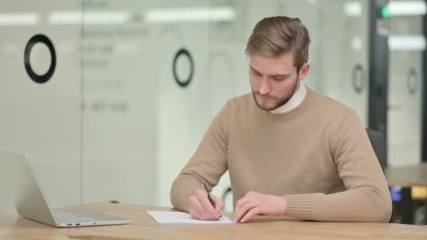 Творческий молодой человек пишет на бумаге в офисе — стоковое видео