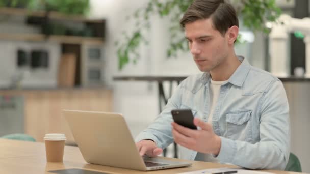 Jovem criativo com laptop usando smartphone no trabalho — Vídeo de Stock