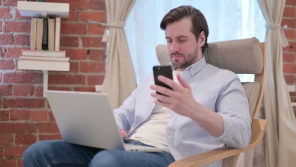 Jovem Casual com Laptop usando Smartphone no Sofá — Vídeo de Stock