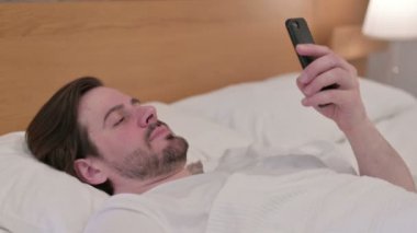 Sıradan genç adam yatakta uyurken akıllı telefon kullanıyor.