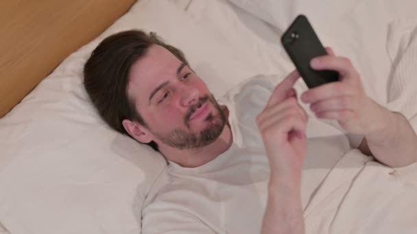ベッドでスマートフォンで成功を収めているカジュアル若い男 — ストック動画