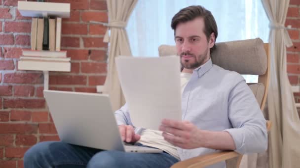 Молода людина з ноутбуком працює над документами на софі — стокове відео
