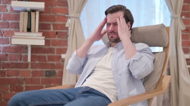 Молодой человек страдает головной болью, сидя на диване — стоковое видео