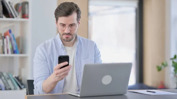 Человек в очках с ноутбуком с помощью смартфона в офисе — стоковое фото