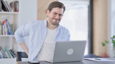 Dizüstü bilgisayarlı, sırt ağrısı olan gözlüklü bir adam. 