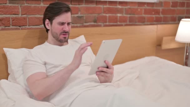 Nenucený mladý muž reagující na ztrátu na tabletu v posteli — Stock video