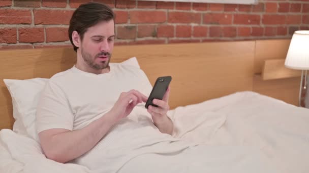 Casual giovane uomo che reagisce alla perdita su smartphone a letto — Video Stock