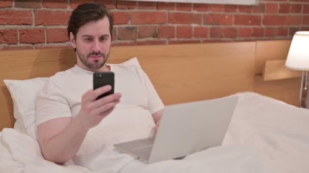 Випадковий молодий чоловік з ноутбуком за допомогою смартфона в ліжку — стокове відео