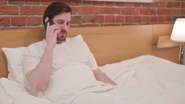 カジュアル若い男｜ベッドの中でスマートフォンで通話中 — ストック動画