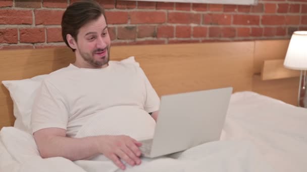 Casual jonge man doet video call op laptop in bed — Stockvideo