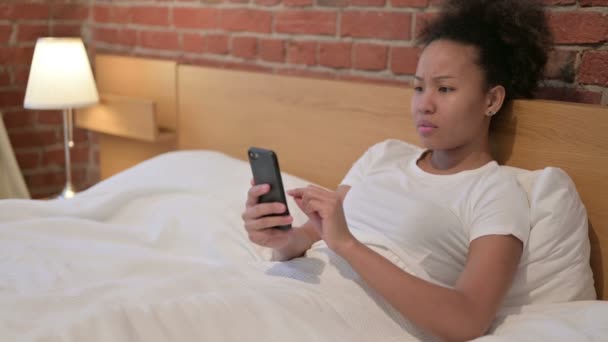 Mujer africana reaccionando a la pérdida en el teléfono inteligente en la cama — Vídeos de Stock