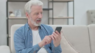 Üst düzey yaşlı adam koltukta Smartphone kullanırken kutlama yapıyor 