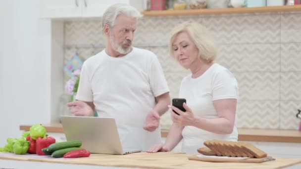 Pareja de ancianos usando en el ordenador portátil y el teléfono inteligente en la cocina — Vídeos de Stock