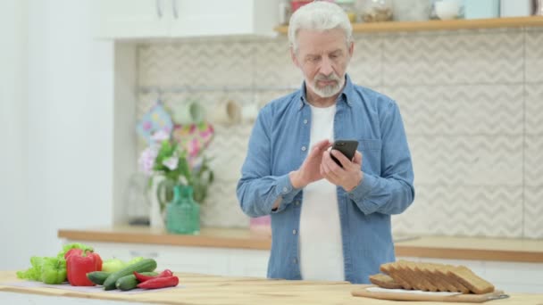 Velho Senior usando Smartphone enquanto corta legumes na cozinha — Vídeo de Stock