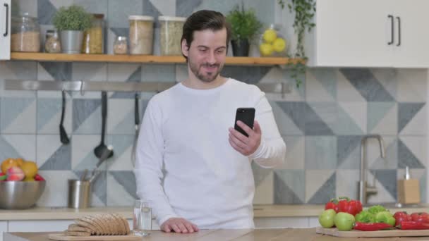 Молодий чоловік робить відеодзвінок на смартфон стоячи на кухні — стокове відео