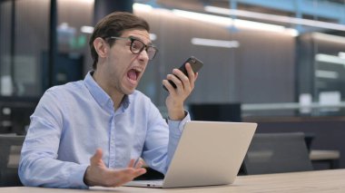 Dizüstü bilgisayarlı kızgın genç adam telefonda konuşuyor.