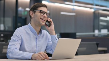 Dizüstü bilgisayarlı genç adam telefonda konuşuyor.