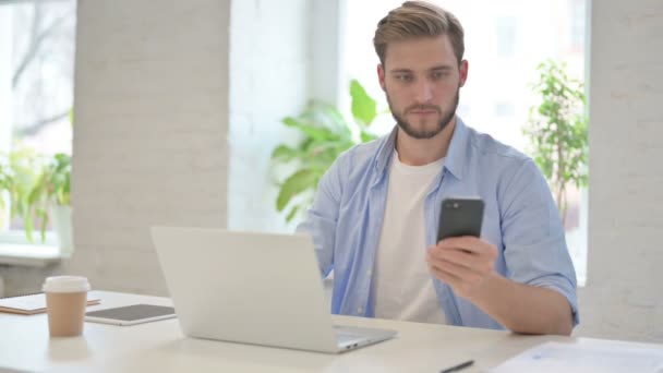 Homem criativo com laptop usando smartphone no escritório moderno — Vídeo de Stock