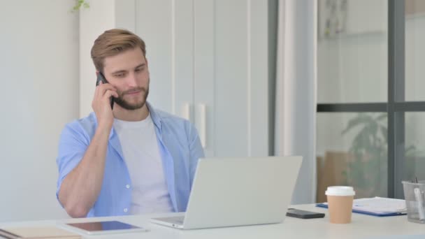 Mladý kreativní muž s notebookem mluví na smartphonu — Stock video