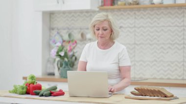 Mutfakta dizüstü bilgisayarda çalışan yaşlı kadın.