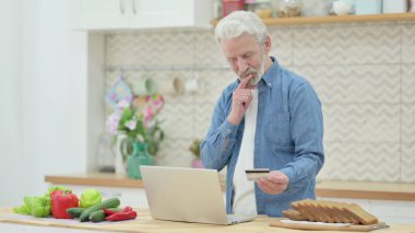 Mutfakta dizüstü bilgisayarda alışveriş yapan yaşlı adam.