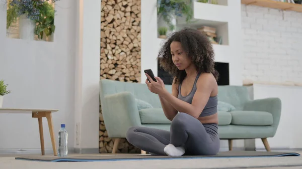 Jovem mulher africana usando Smartphone em excercise Mat em casa — Fotografia de Stock