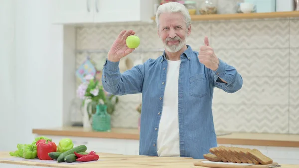 Old Man Showing Thumbs up while Holding Apple in Kitchen