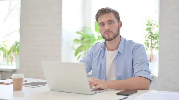 Junger Mann blickt in modernem Büro mit Laptop auf Kamera — Stockfoto