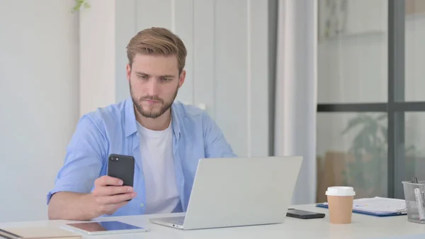 Jonge man met laptop met behulp van smartphone op het werk — Stockfoto