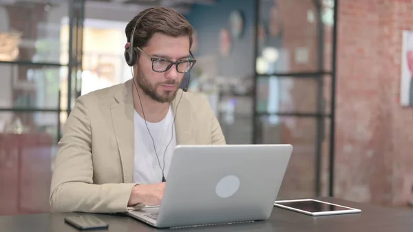 Uomo che indossa auricolare di lavoro sul computer portatile al lavoro — Foto Stock