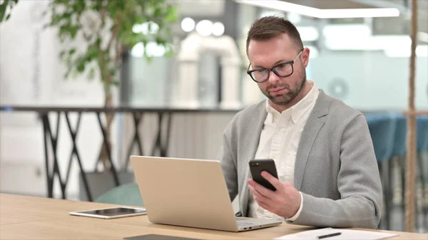 Кавказский молодой человек просматривает Интернет на смартфоне и ноутбуке в офисе — стоковое фото