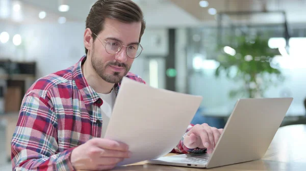 Grave giovane uomo che lavora sul computer portatile con documenti in Cafe — Foto Stock