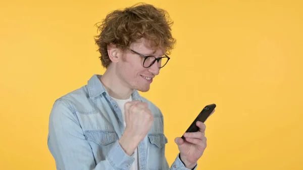 Рыжий молодой человек празднует на смартфоне на жёлтом фоне — стоковое фото
